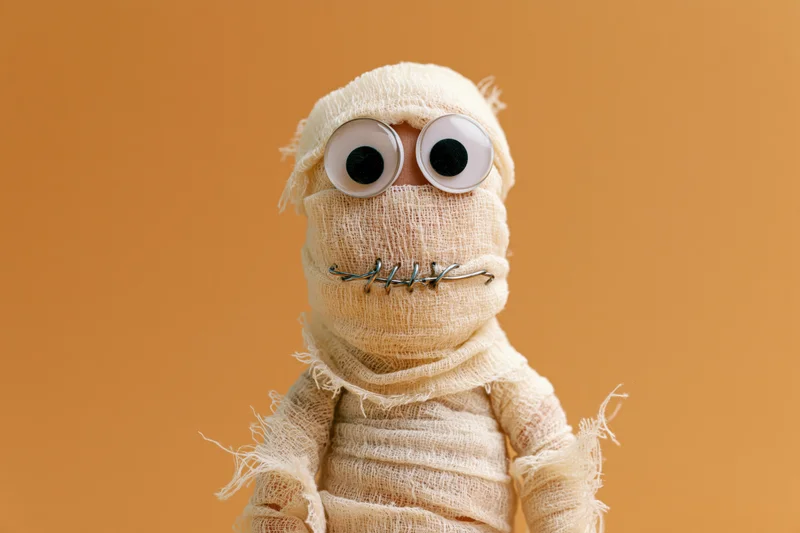 Handmade mummy figurine wrapped in cream gauze with googly eyes and wire mouth against orange background