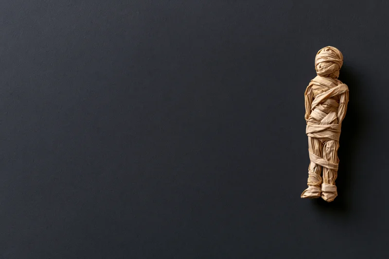 Handmade mummy figurine wrapped in tan paper on dark surface