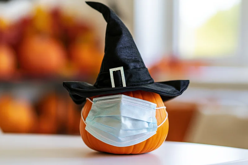 Small orange pumpkin wearing black witch hat and blue medical mask on white table with blurred pumpkins in background