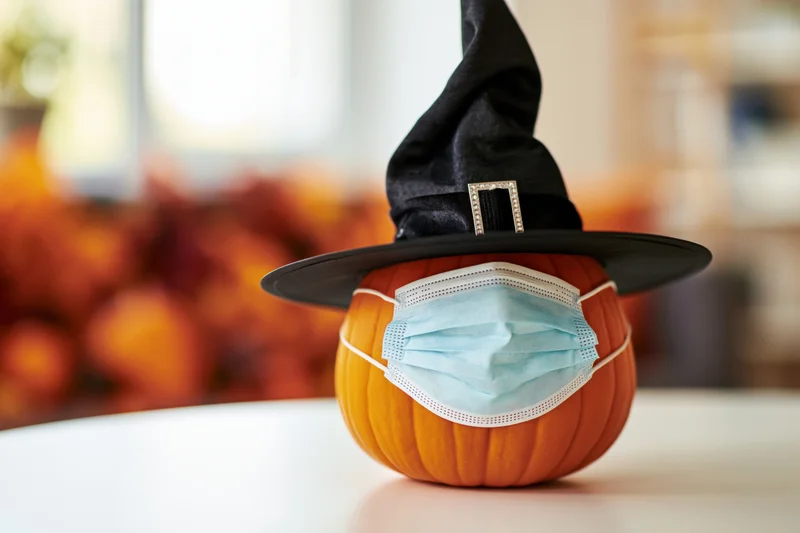 Small orange pumpkin wearing black witch hat and blue face mask on white table