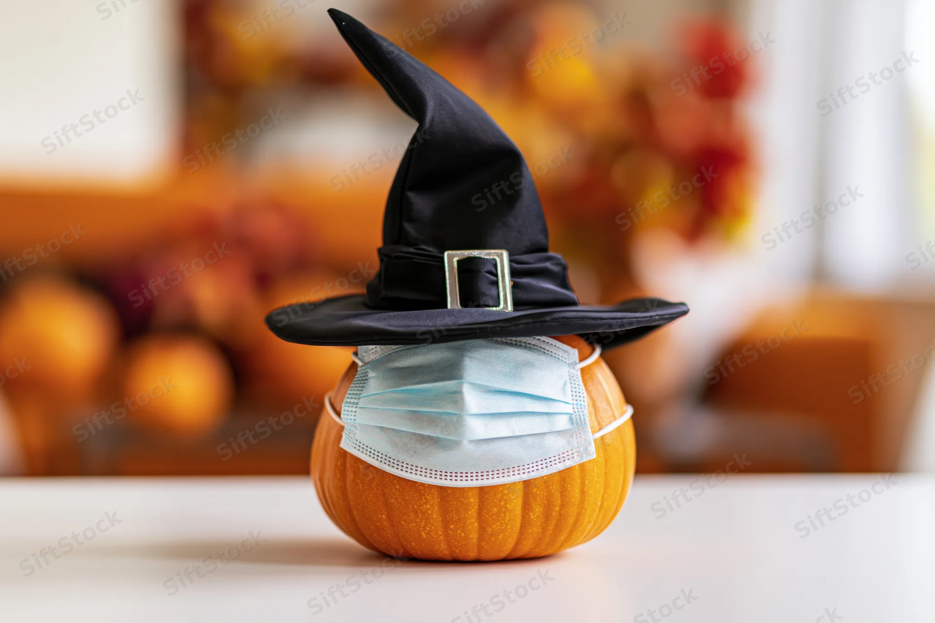 Small orange pumpkin wearing black witch hat and face mask on white table