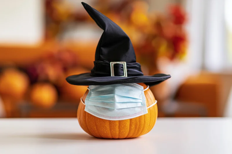 Small orange pumpkin wearing black witch hat and face mask on white table