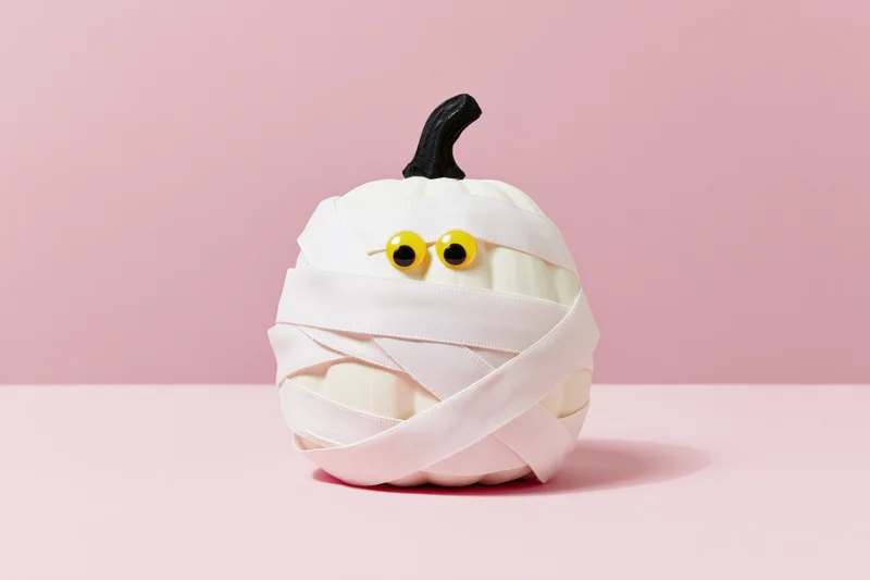 White pumpkin wrapped in white ribbon with yellow eyes and black stem on pink surface