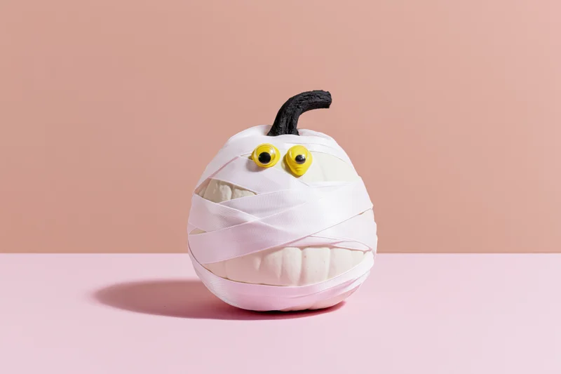 White pumpkin wrapped in ribbon as a mummy decoration on a pink surface