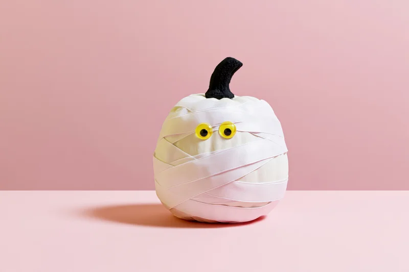 White pumpkin wrapped in ribbon with googly eyes and black stem on pink background