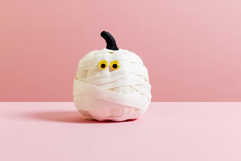 White pumpkin wrapped in ribbon with googly eyes as a mummy decoration on pink surface