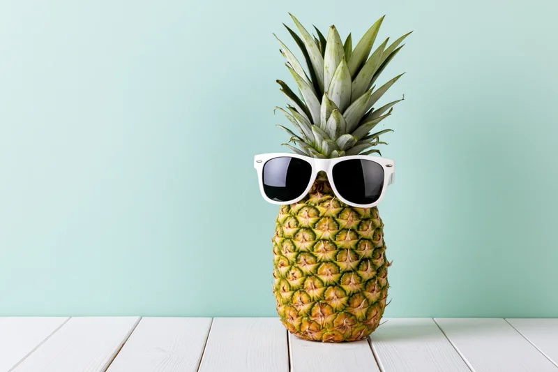 Pineapple wearing white-framed sunglasses on a white wooden table against a mint green background