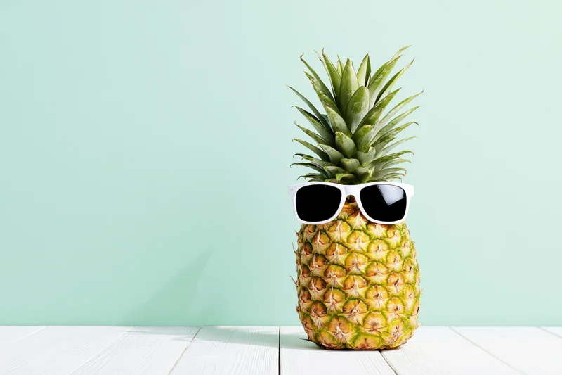 Pineapple wearing white-framed sunglasses on a white wooden table against a mint background