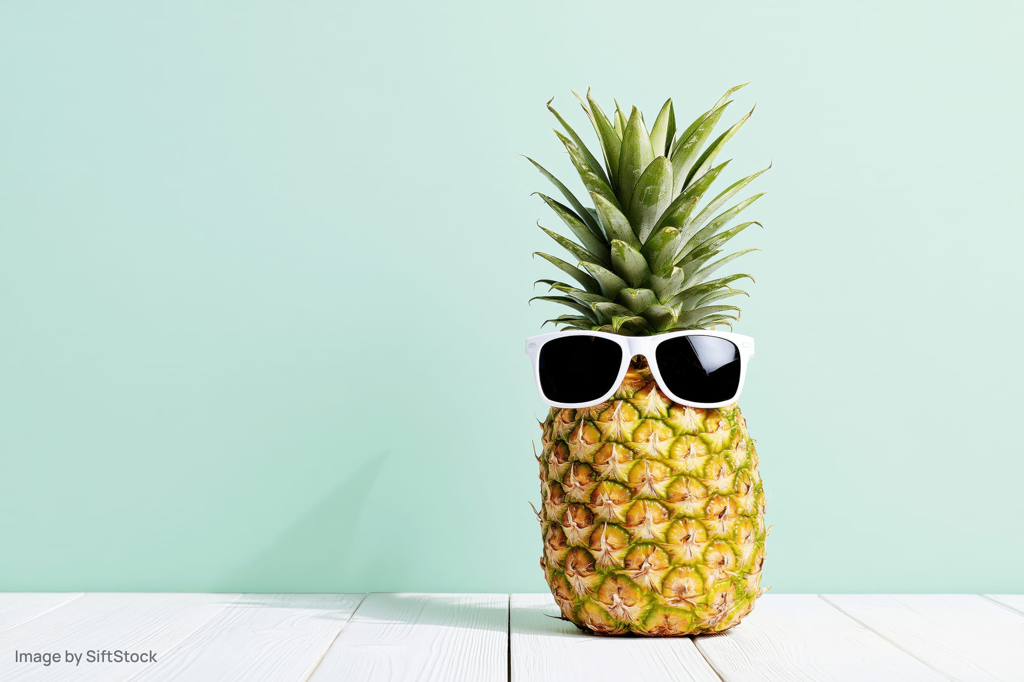Pineapple wearing white-framed sunglasses on a white wooden table against a mint background