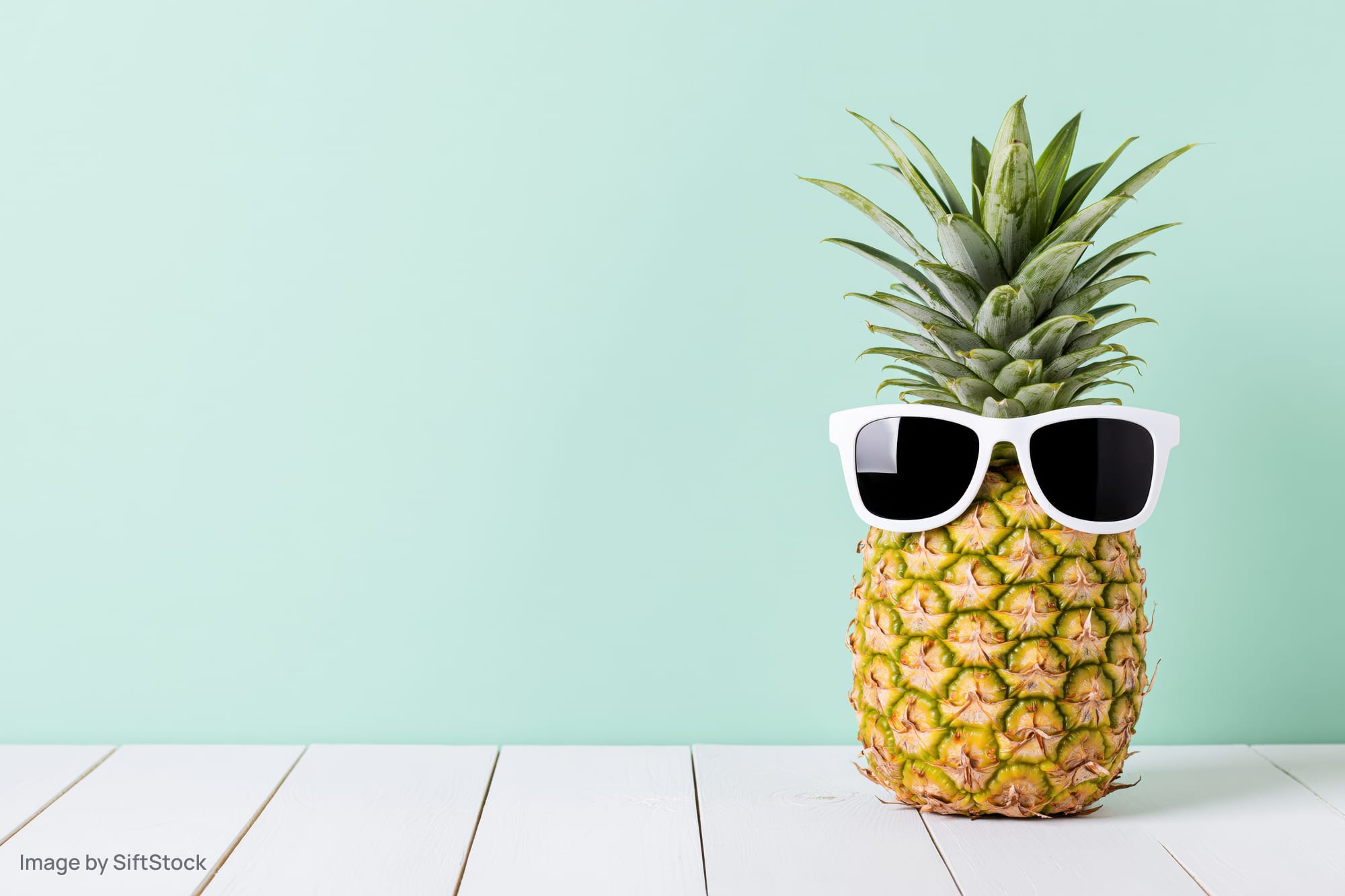 Pineapple wearing white sunglasses on a white wooden table against a mint green background