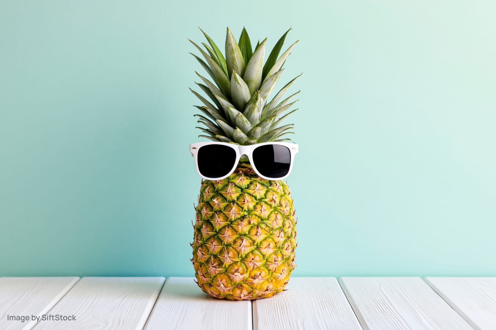 Pineapple wearing white-framed sunglasses on a white wooden table against a mint green background