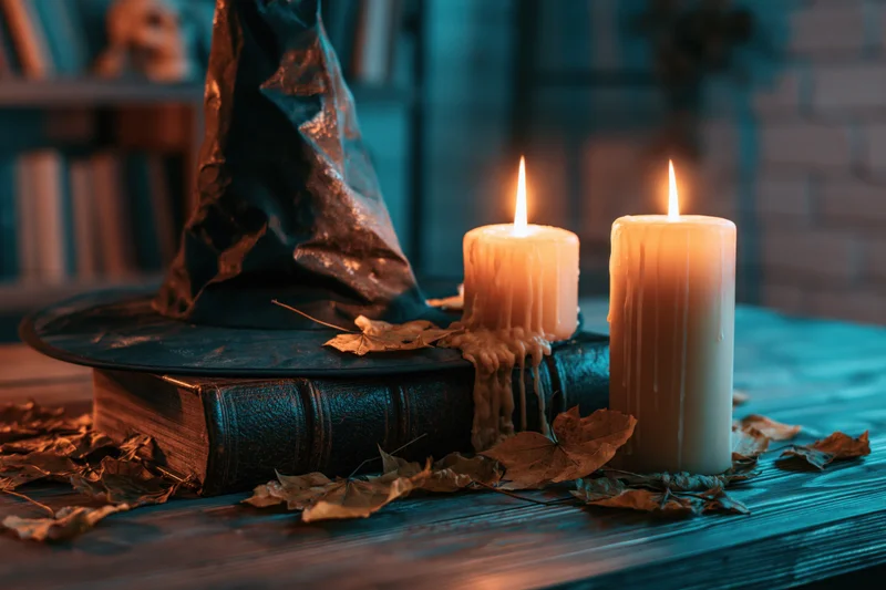 Witch hat resting on an old leather-bound book beside two lit candles on a wooden table scattered with dried autumn leaves