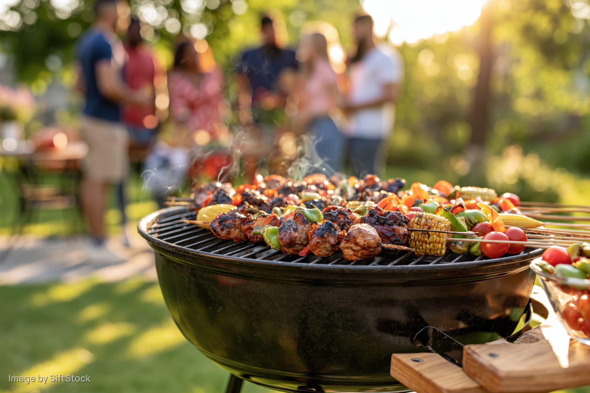 Charcoal grill with skewered meats and vegetables cooking outdoors during a summer gathering in a backyard garden