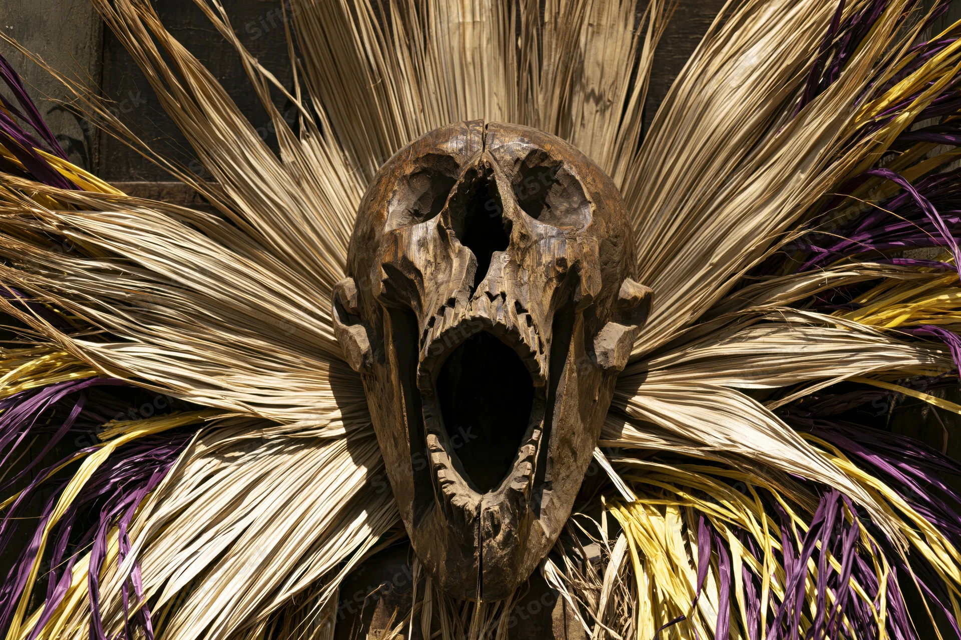 Carved wooden skull mask with open screaming mouth surrounded by radiating dried palm fronds in purple and yellow