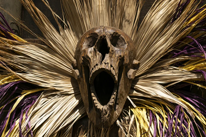 Carved wooden skull mask with open screaming mouth surrounded by radiating dried palm fronds in purple and yellow