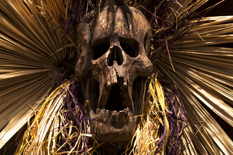 Carved wooden skull mask with open screaming mouth surrounded by dried palm fronds and purple straw