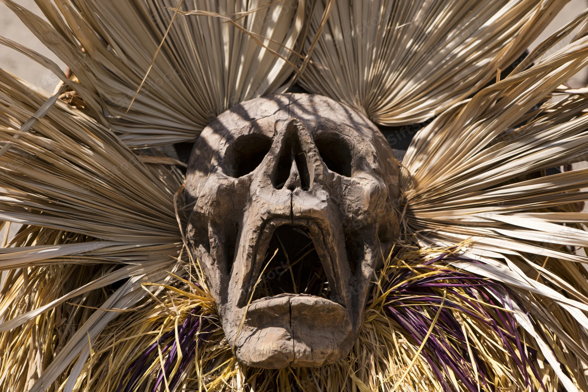 Carved wooden skull mask with open screaming mouth surrounded by dried palm fronds