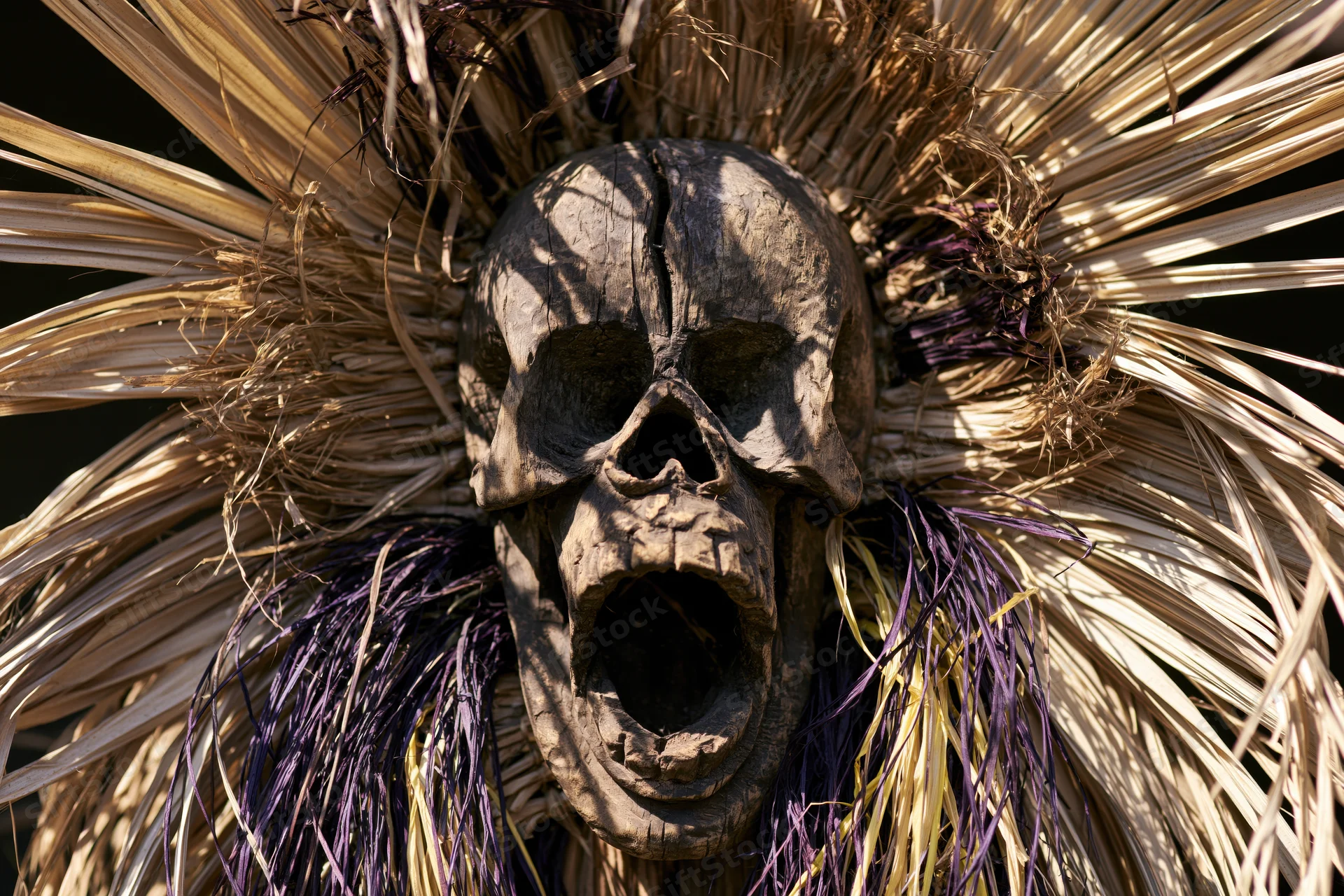 Carved wooden skull mask with open screaming mouth surrounded by dried palm fronds