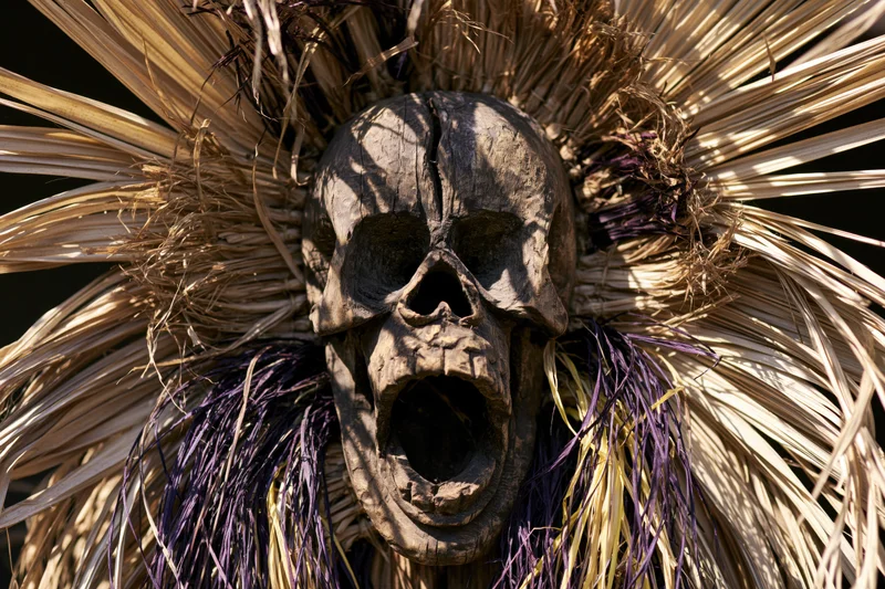 Carved wooden skull mask with open screaming mouth surrounded by dried palm fronds