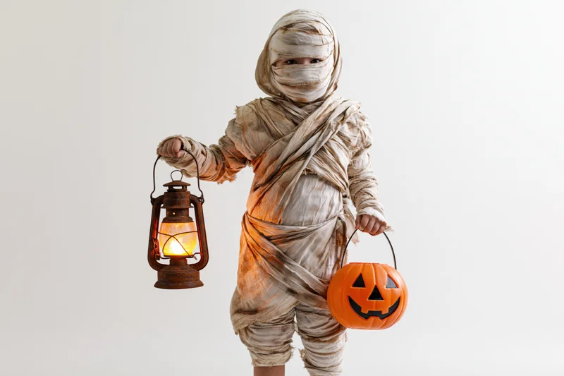 Child in mummy costume holding lantern and pumpkin bucket in studio