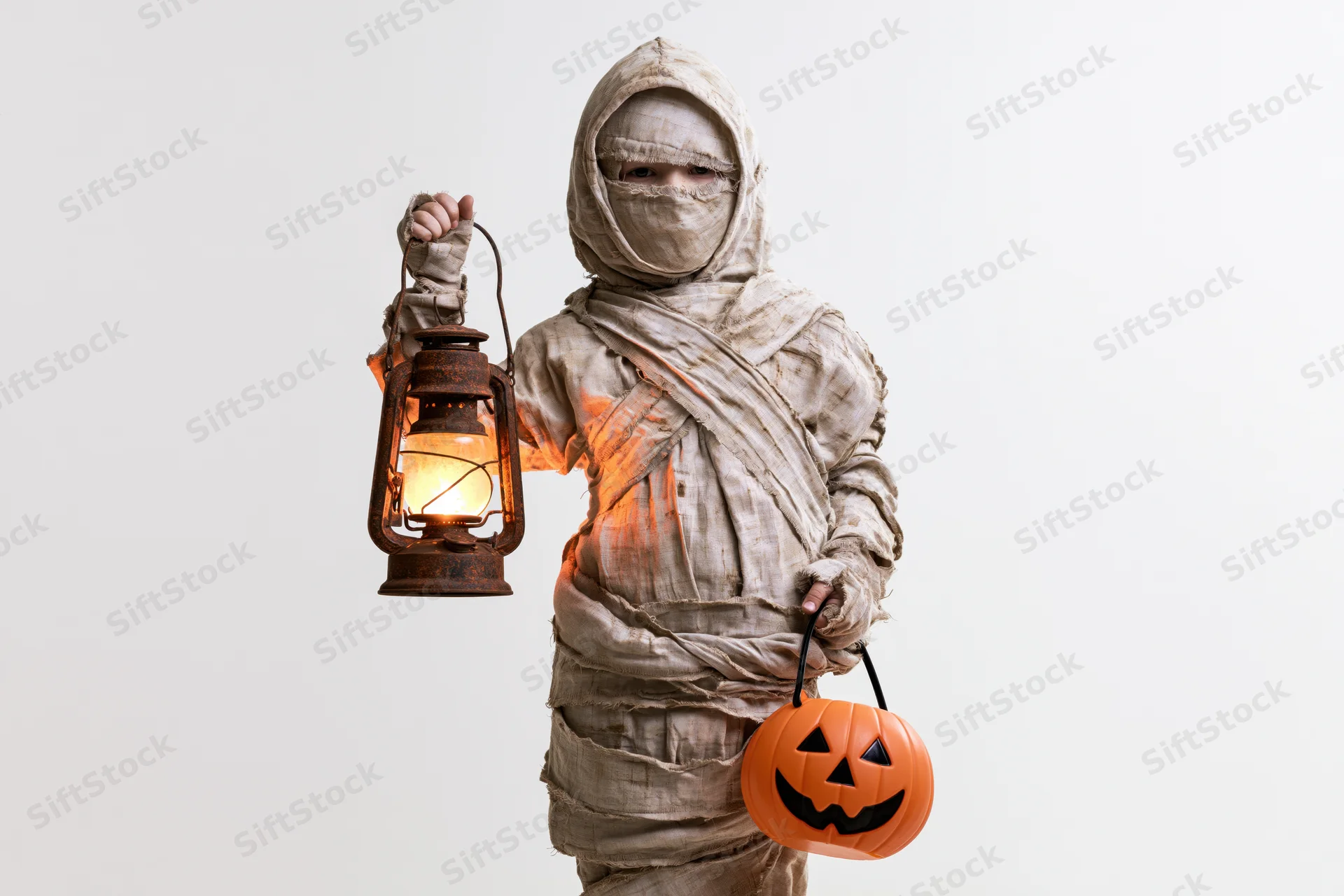 Child in mummy costume holding lantern and pumpkin bucket against white background