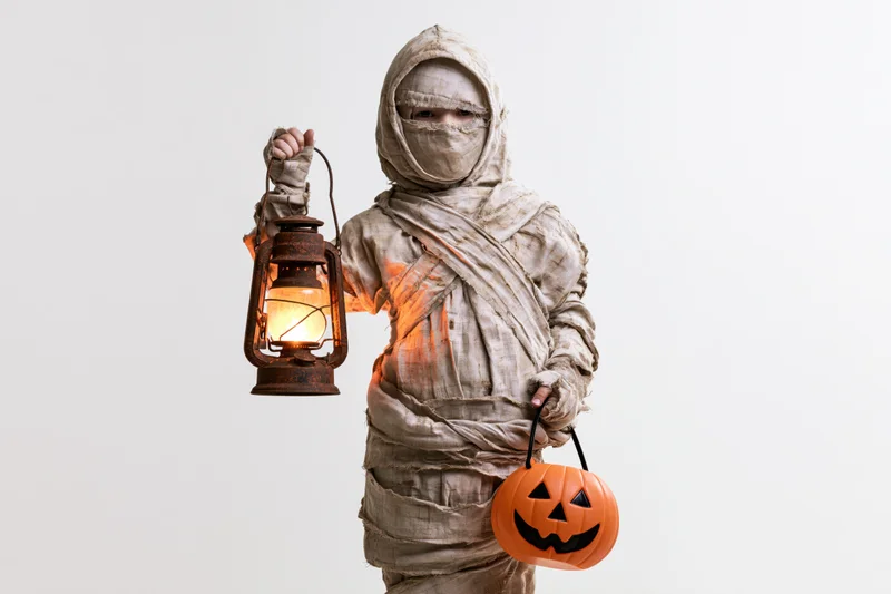 Child in mummy costume holding lantern and pumpkin bucket against white background
