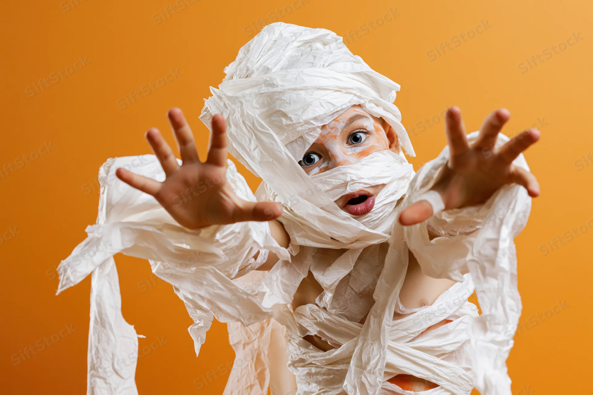 Child in homemade mummy costume with paper wrappings posing playfully against orange background