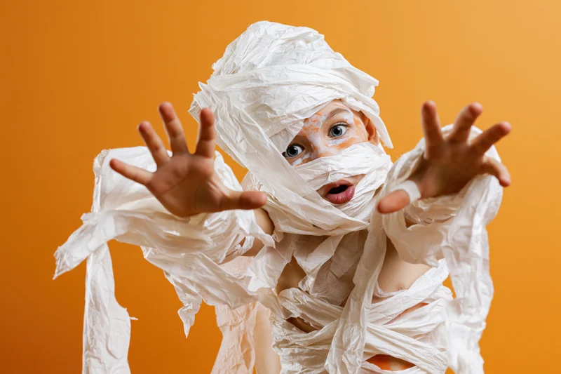 Child in homemade mummy costume with paper wrappings posing playfully against orange background