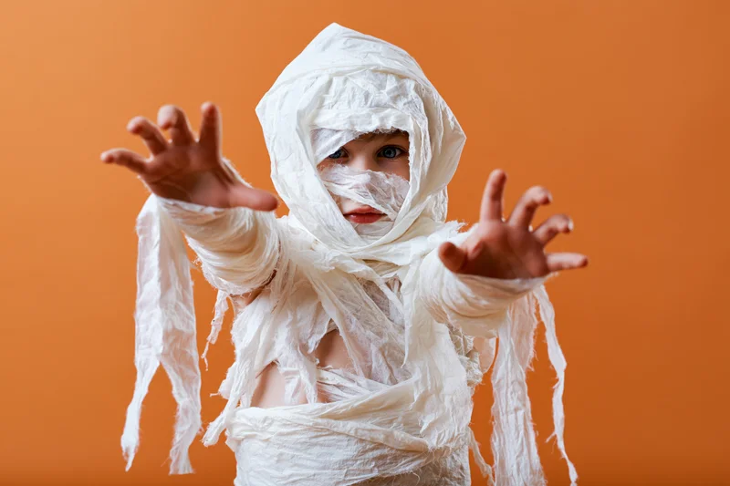Child in homemade mummy costume with arms outstretched against orange background
