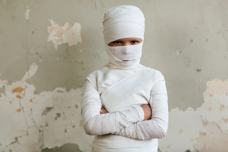 Child wrapped in white gauze bandages standing with arms crossed in front of peeling wall