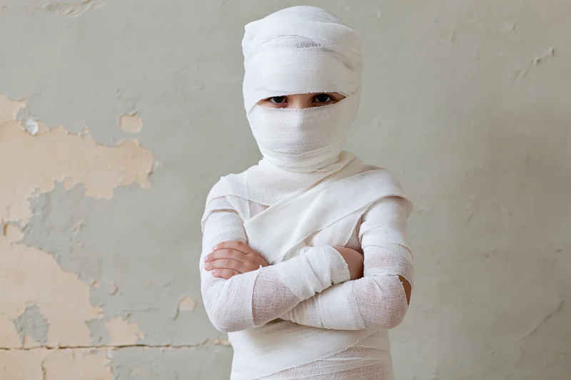 Child wrapped in white gauze bandages with arms crossed against a peeling wall