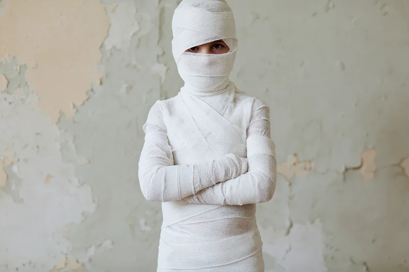 Child wrapped in white medical bandages with arms crossed against peeling wall