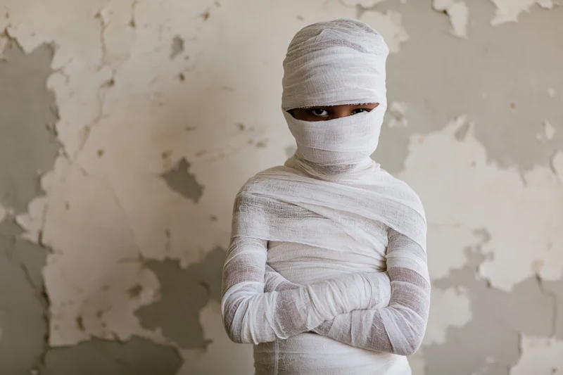 Child wrapped in white gauze bandages with arms crossed standing before peeling wall