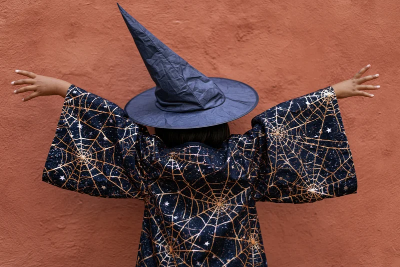 Child in witch costume with arms outstretched against orange wall