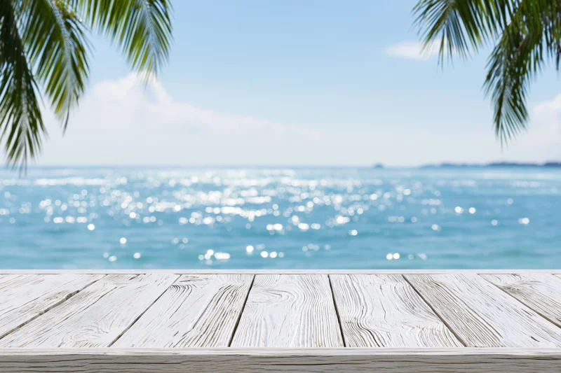 Weathered white wooden table in front of a tropical beach with ocean and palm leaves