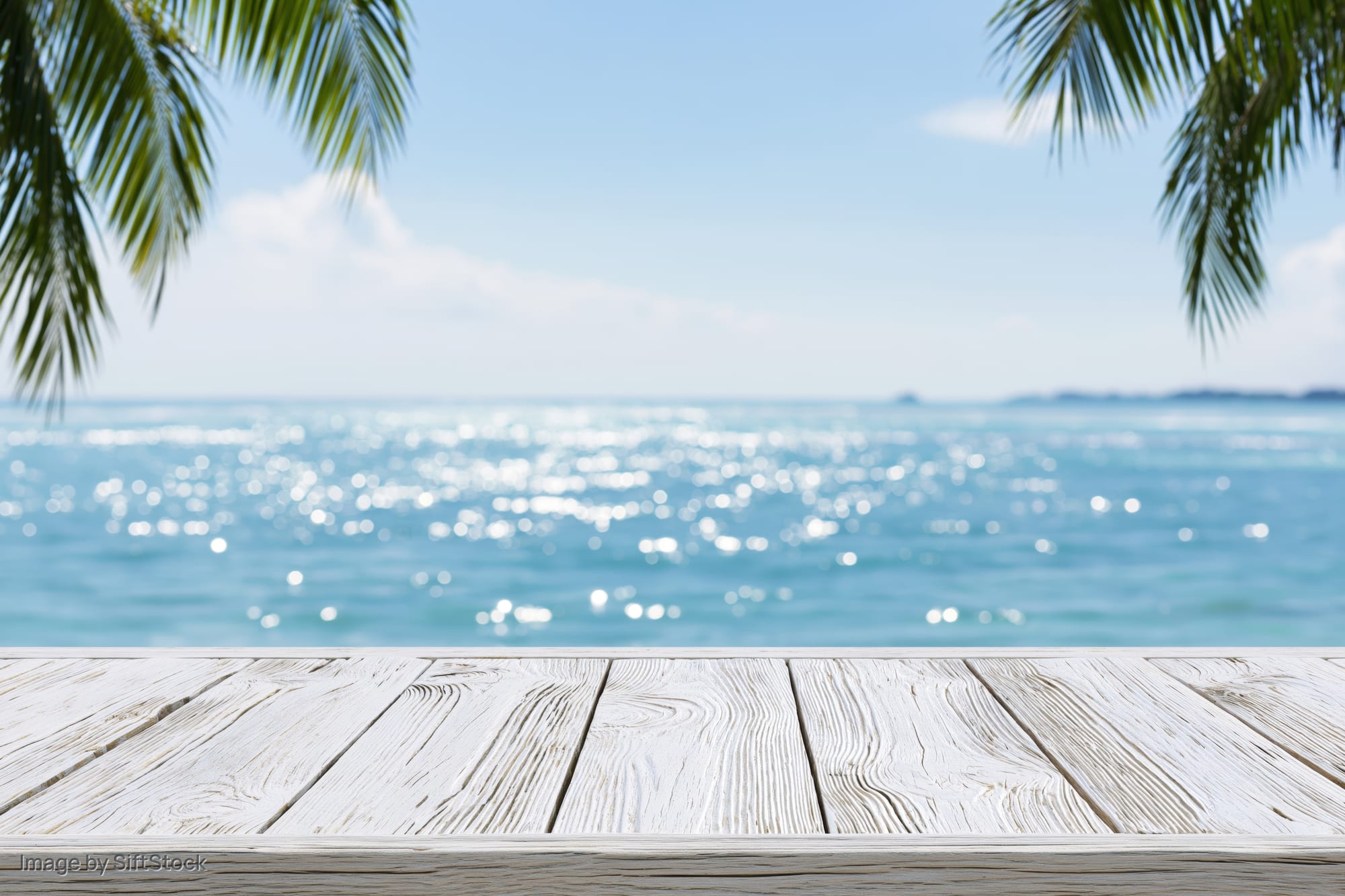 Weathered white wooden table in front of a tropical beach with ocean and palm leaves