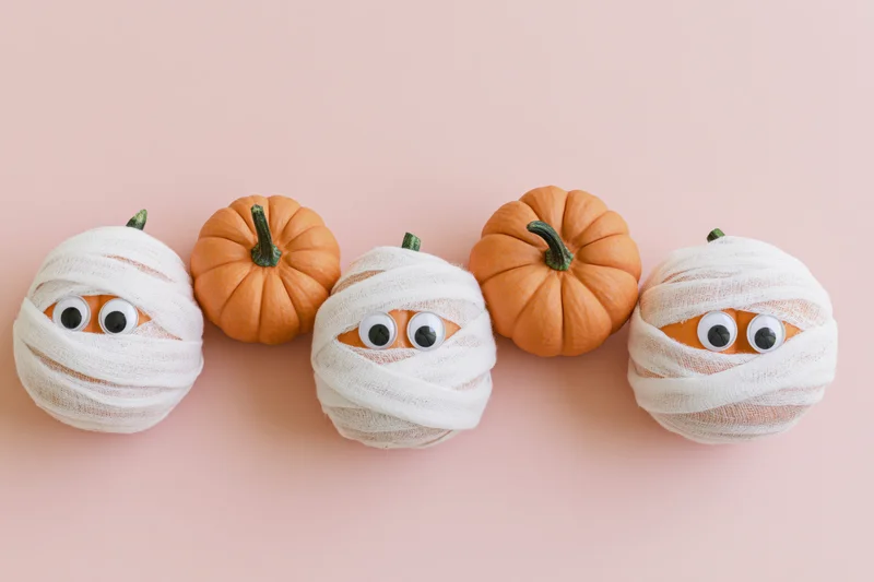Four small pumpkins wrapped in white gauze with googly eyes arranged in a row on a pink background