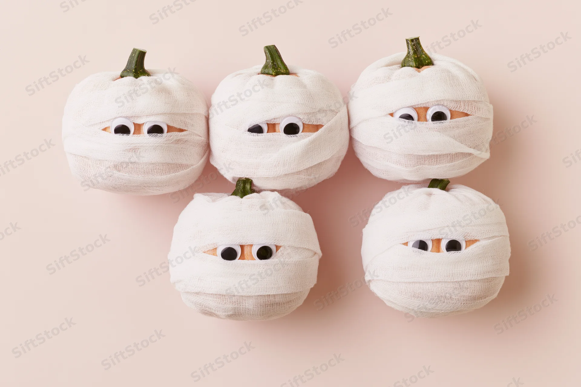 Five small pumpkins wrapped in white gauze bandages with googly eyes on a plain background