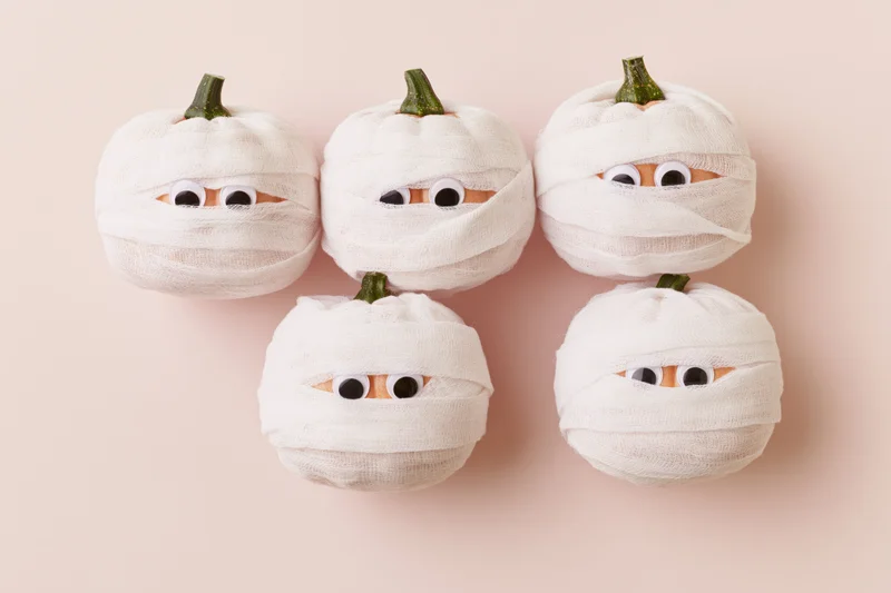 Five small pumpkins wrapped in white gauze bandages with googly eyes on a plain background