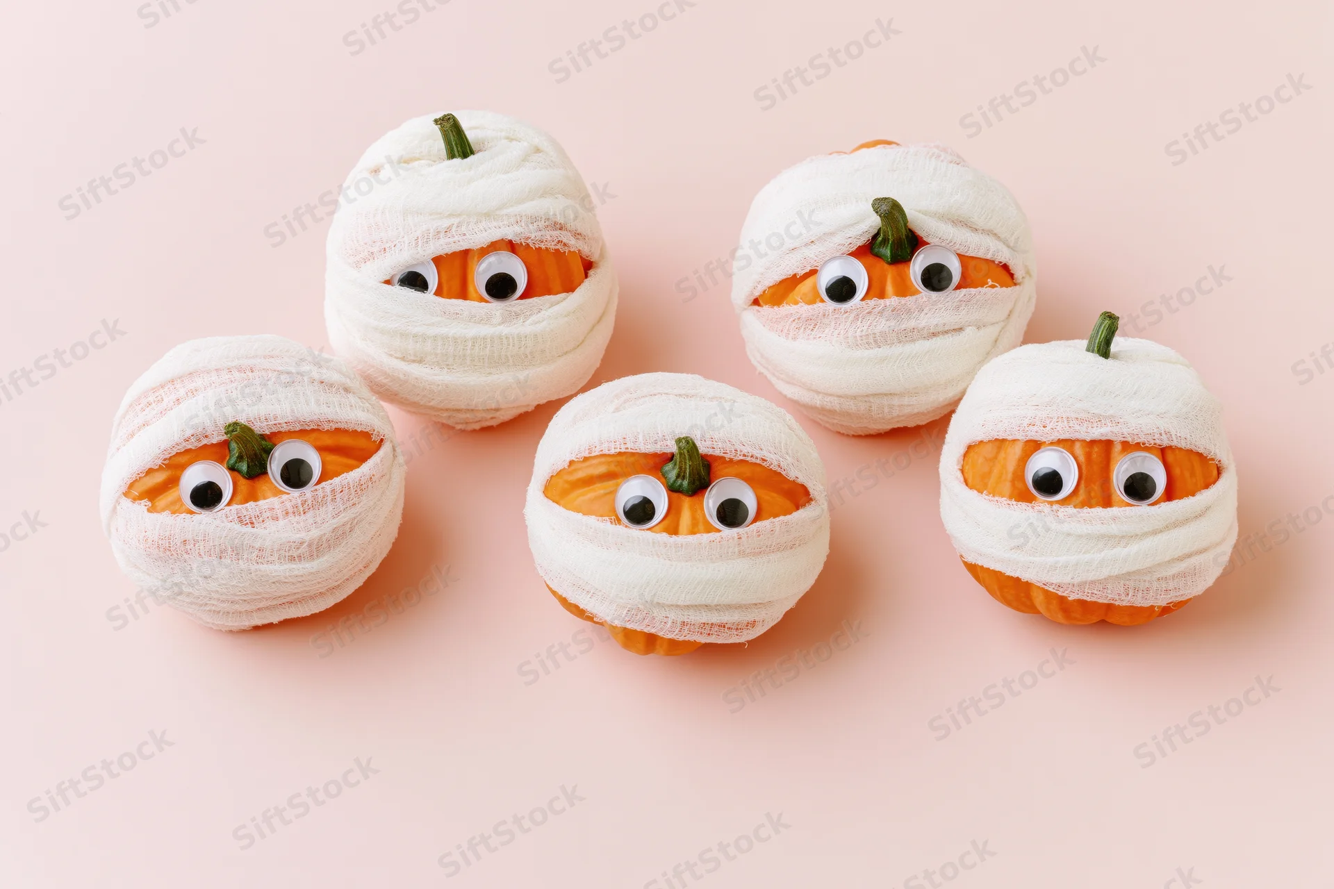 Five small orange pumpkins wrapped in white gauze bandages with googly eyes on a pink background