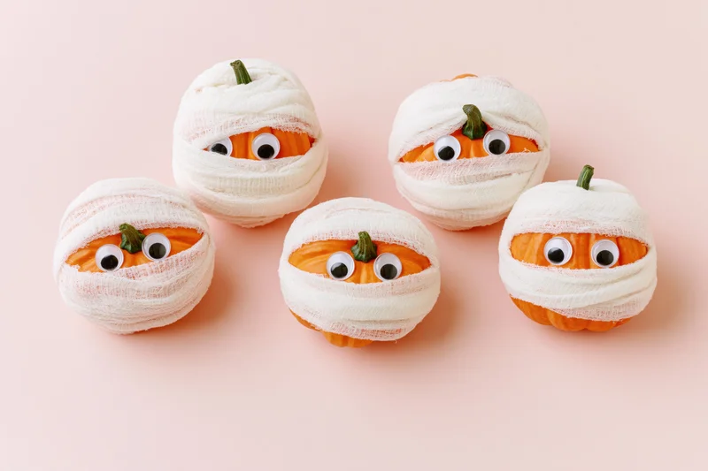 Five small orange pumpkins wrapped in white gauze bandages with googly eyes on a pink background