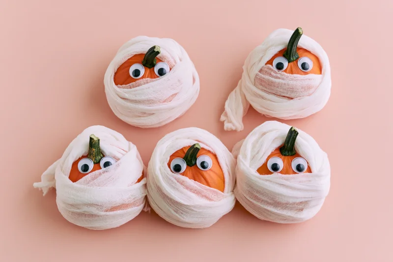 Five small orange pumpkins wrapped in white gauze with googly eyes arranged on a pink surface
