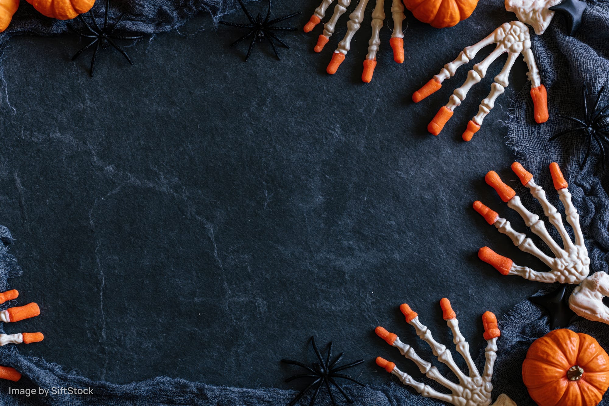 Halloween flat lay with skeleton hands, pumpkins and spiders arranged on dark slate stone surface with copyspace