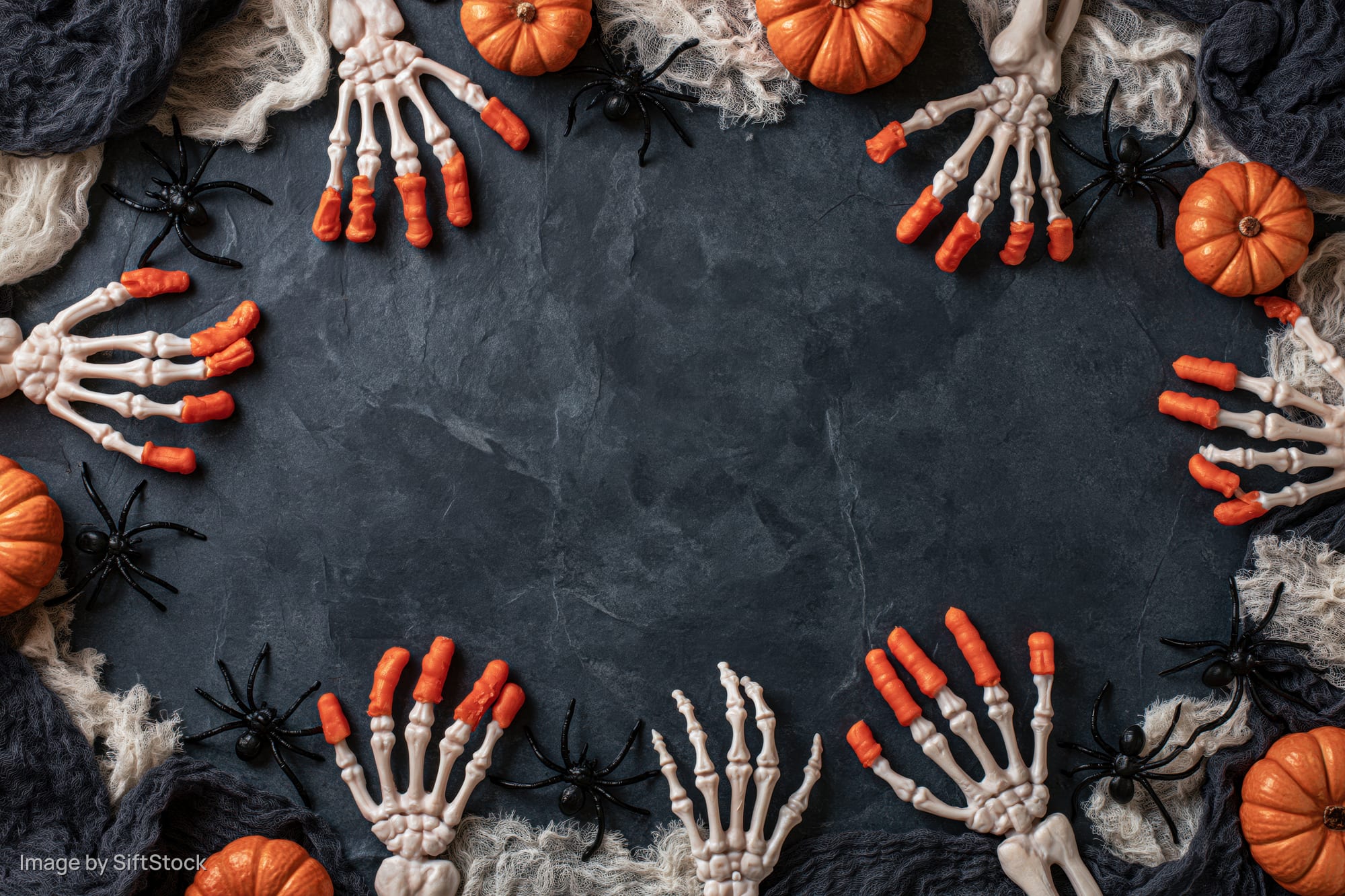 Halloween flat lay with skeleton hands, pumpkins and spiders arranged in a frame on dark slate stone surface