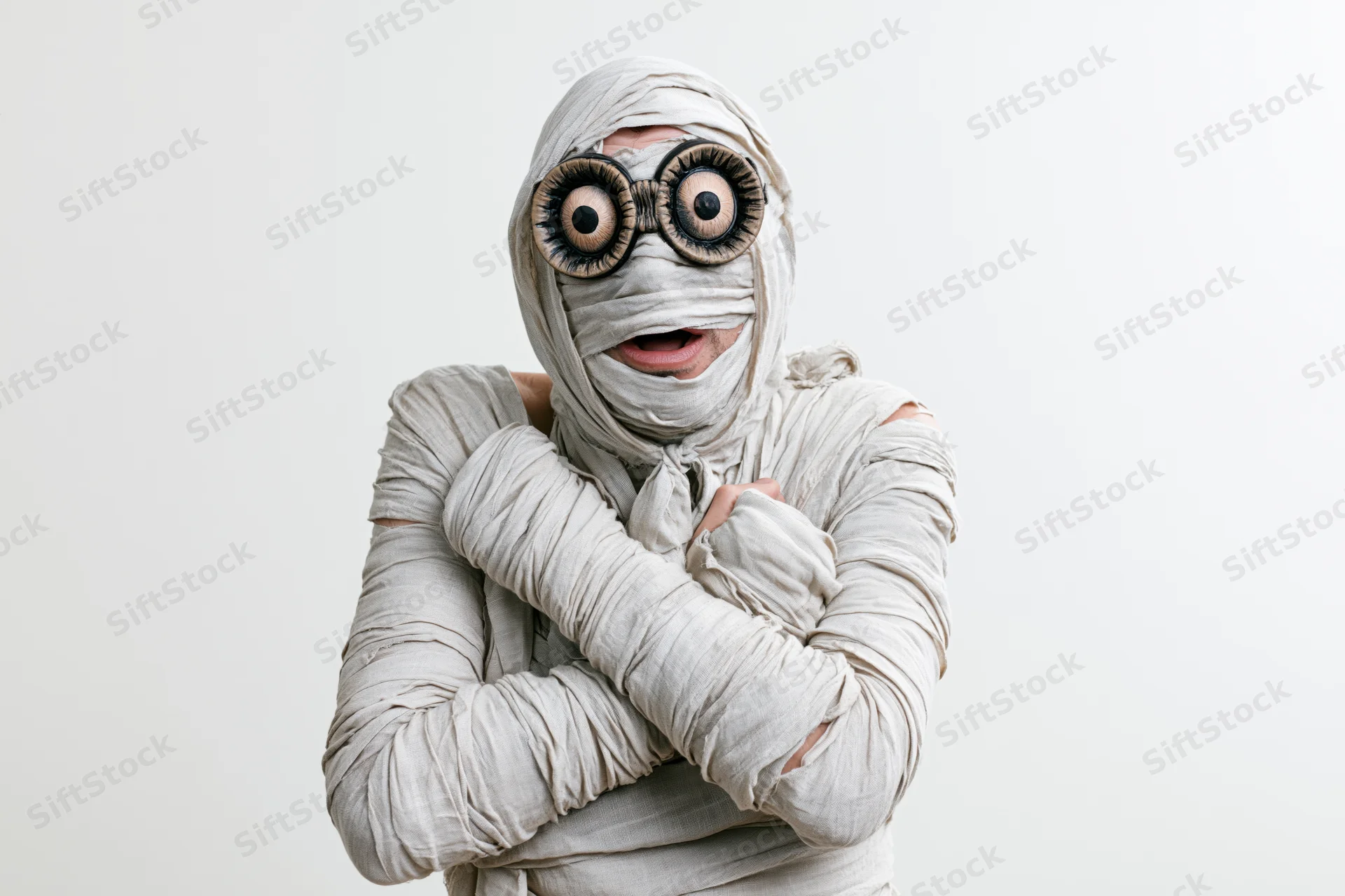 Man dressed as halloween mummy with oversized eyes and crossed arms against white background