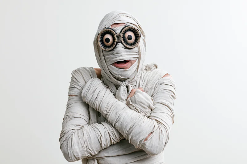 Man dressed as halloween mummy with oversized eyes and crossed arms against white background