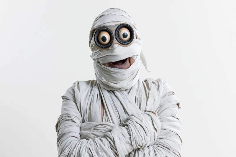 Man dressed as halloween mummy with arms crossed and large eyes smiling at camera in studio