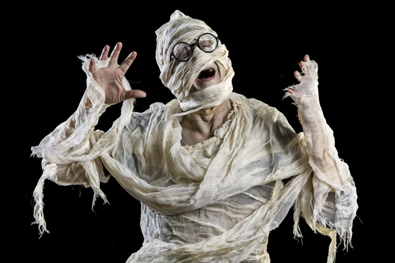 Man in homemade mummy costume laughing with arms raised against black background
