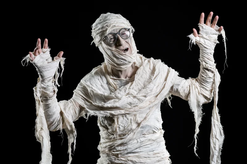 Man in homemade mummy costume laughing with arms raised against black background