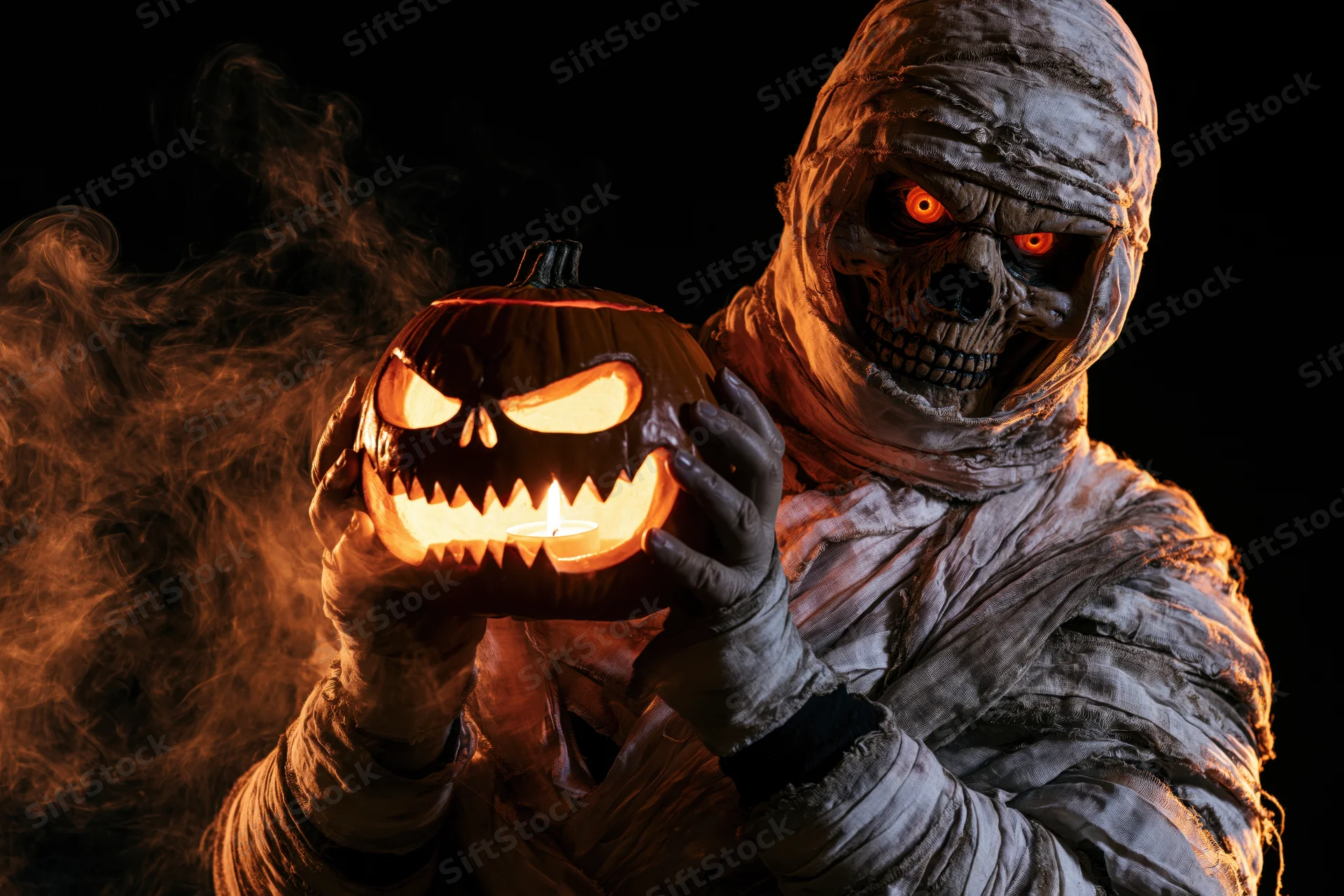 Mummy in bandages holding a carved pumpkin with glowing eyes and smoke in dark setting